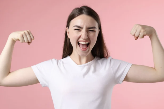 Junge Frau mit langen Haaren, die lächelt und ihre starken Arme zeigt, vor rosa Hintergrund.