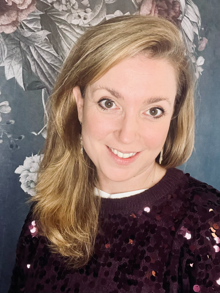 Frau mit blonden Haaren und schimmerndem Oberteil vor floralem Hintergrund.