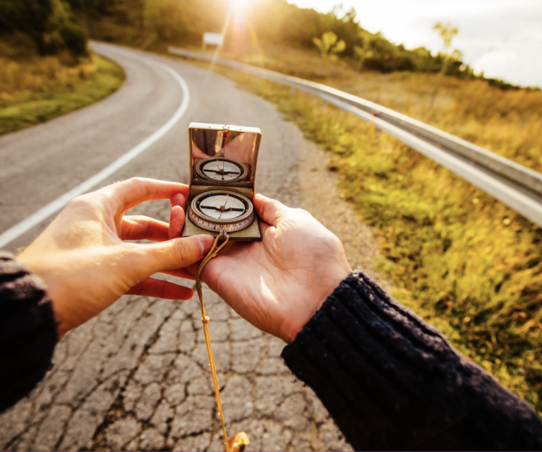 Eine Person hält einen Kompass auf einer kurvenreichen Landstraße in der Abendsonne.